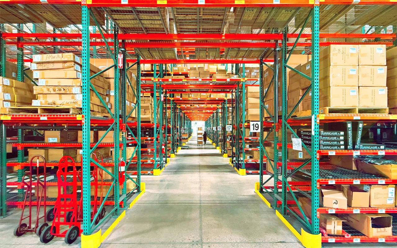 A long warehouse aisle between green and orange pallet racks stacked with cartons